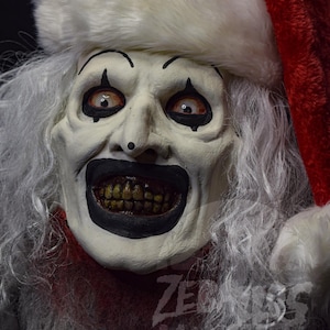 Puede incluir: Una máscara de Papá Noel espeluznante blanca y roja con una amplia sonrisa de dientes y ojos negros. La máscara tiene una barba blanca y un gorro de Papá Noel rojo.