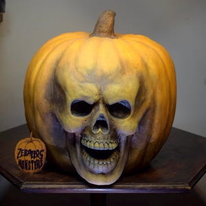 Peut inclure: Une grande sculpture de citrouille jaune-orange, ronde, présentant un visage de crâne humain sculpté avec une bouche ouverte et des dents visibles. Une petite étiquette ronde en bois avec le texte ZEBALLOS MONSTERS en violet est à gauche.