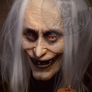 Puede incluir: Un rostro detallado y envejecido con cabello blanco y una sonrisa siniestra. La cara tiene arrugas profundas, cuencas oscuras y una nariz prominente. Una pequeña calabaza con el texto "Zebatos Monsters" está en primer plano.