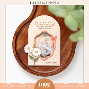 Peut inclure: Un cadre photo en bois avec un motif de marguerite rose et blanche. Le cadre est conçu pour accueillir une photo de 2,5x3,5 pouces. Le texte "mommy" est écrit en cursive avec les mots "You make everyday bloom with love" en dessous. Le cadre comprend également le texte "Winnie & Callum".