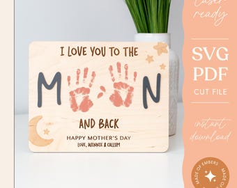 May include: A wooden sign with the text "I love you to the moon and back" and two handprints in pink. The sign also includes a crescent moon and stars. The text "Happy Mother's Day" and "Love, Winnie & Callum" is below the handprints.