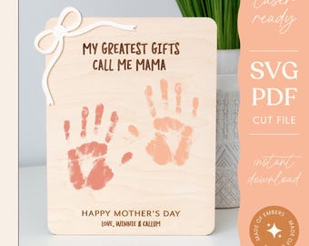Op de afbeelding: Een houten bord met de tekst "Mijn grootste geschenken noemen me mama" en twee handafdrukken in roze en oranje. Het bord heeft ook de tekst "Gelukkige Moederdag" en "Lieve groet, Winnie & Callum".