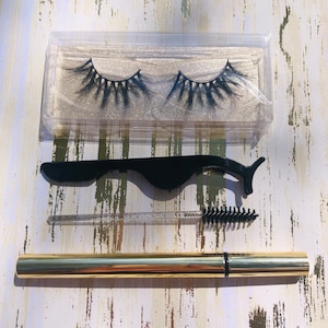 Könnte beinhalten: Ein Satz künstlicher Wimpern in einem durchsichtigen Plastikbehälter, ein schwarzer Wimpernapplikator, ein transparenter Applikator mit einem schwarzen Wimpernbürstchen und ein goldener Eyeliner-Stift.