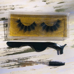 May include: A set of false eyelashes in a clear plastic case with a black lash applicator and a black lash brush. The eyelashes are in a clear plastic case with a gold glitter background.