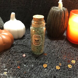 Pode incluir: Uma pequena garrafa de vidro cheia de cristais verdes, rotulada como "POISON" com uma caveira. A garrafa tem uma rolha de cortiça e é cercada por decorações de Halloween, incluindo abóboras e uma vela.