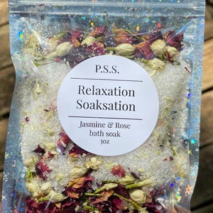 May include: A clear plastic bag filled with white bath salts, dried rose petals, and jasmine flowers. The bag has a label that reads "P.S.S. Relaxation Soaksation Jasmine & Rose bath soak 3 oz."