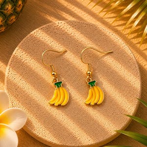 Peut inclure: Une paire de boucles d'oreilles dorées avec un motif de banane. Chaque boucle d'oreille présente une grappe de bananes jaunes avec des sommets verts. Les boucles d'oreilles sont sur une surface ronde et beige avec un thème tropical.