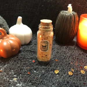 Pode incluir: Uma pequena garrafa de vidro com rolha, rotulada "POISON" com uma caveira. Contém grânulos laranja e castanhos. O fundo inclui abóboras decorativas e uma vela acesa, criando um ambiente de Halloween.