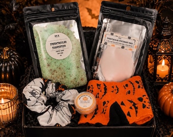 May include: A gift box containing a black pouch of green bath soak labeled Spooktacular Soaksation, a black pouch of stationery, a white scrunchie with black spiderweb patterns, a small orange jar, and orange socks with black jack-o'-lantern faces.