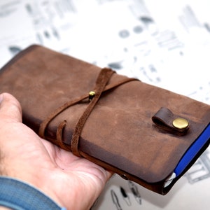 May include: A brown leather journal with a strap closure and a brass button. The journal is tied with a brown leather strap and has a distressed look.