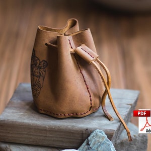 May include: A brown leather drawstring pouch with a stitched design and a black and white image of a person. The pouch has a long brown leather strap.