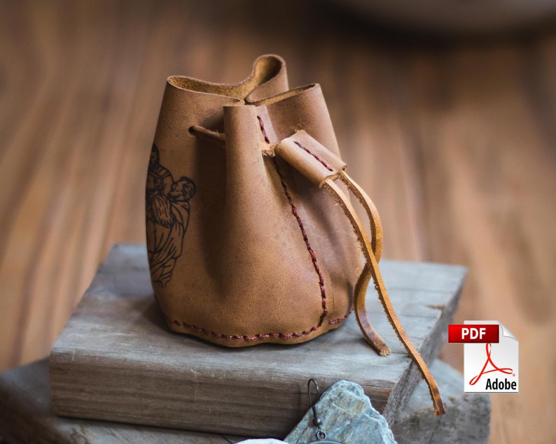 May include: A brown leather drawstring pouch with a stitched design and a black and white image of a person. The pouch has a long brown leather strap.