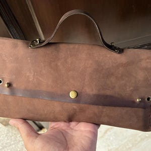 May include: A brown leather clutch bag with a top handle and brass hardware. The bag has a flap closure and a single compartment.