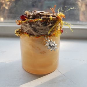 May include: A small glass jar with a brown lid decorated with twine, yellow and orange string, and red beads. A silver charm in the shape of a sun hangs from the jar.