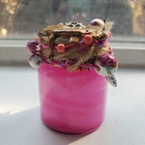Puede incluir: Un pequeño frasco de vidrio rosa con una tapa dorada y una variedad de cuentas y abalorios coloridos alrededor de la tapa.