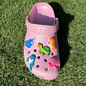 Puede incluir: Crocs rosas con abalorios Jibbitz de criaturas marinas. Los abalorios incluyen una raya azul, una tortuga marina verde, un pulpo rosa, un delfín azul y un caballito de mar naranja.