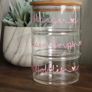 Op de afbeelding: Drie glazen potten met bamboe deksels, gestapeld op elkaar. De potten zijn gelabeld met roze letters en hartjes: "Hair Changer", "Hair Grips" en "Bottles".