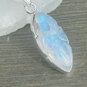 May include: A silver pendant with a carved moonstone. The moonstone is oval shaped and has a blue iridescent sheen.