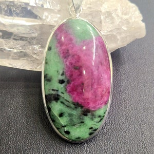 Puede incluir: Un colgante de plata con una piedra ovalada verde y rosa con motas negras. El colgante está engastado en un bisel de plata.
