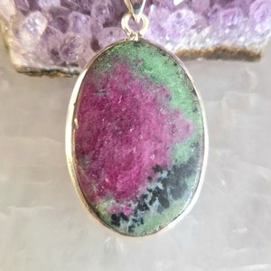 Puede incluir: Colgante ovalado con una fianza plateada, con una piedra de rubí zoisita. La piedra muestra una mezcla de tonos rosa intenso, verde y negro. El colgante está sobre una superficie blanca suave, con un racimo de cristal púrpura en el fondo.