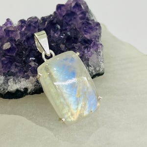 Pode incluir: Um pingente de prata com uma pedra de lua retangular iridescente com um brilho azul e branco. O pingente está colocado sobre um fundo de cristais de ametista púrpura.