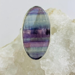 Può includere: Anello ovale con una pietra di fluorite lucidata. La pietra presenta bande di colori viola, lavanda, blu e trasparenti, incastonata in una fascia di metallo color argento. Il design dell'anello è semplice, evidenziando la bellezza naturale della gemma.