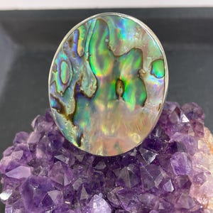 May include: A large, round ring with an iridescent abalone shell centre, set in a silver band. The shell displays a mix of green, blue, and gold hues. The ring rests on a cluster of purple amethyst crystals.