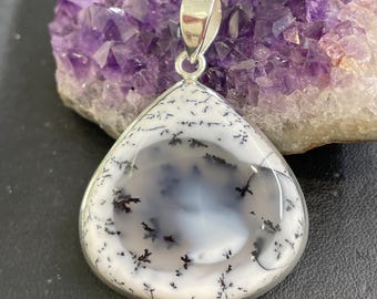 Op de afbeelding: Een druppelvormige hanger met een zilveren beugel. De hanger heeft een witte en grijze dendrietagaatsteen met zwarte insluitsels, gezet in een zilveren bezel. Op de achtergrond een paars amethistkristal.