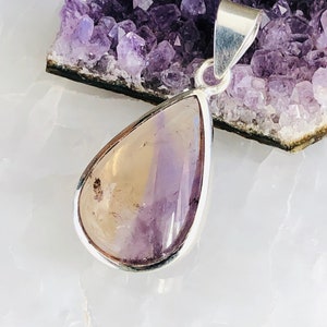 Puede incluir: Un colgante en forma de l&aacute;grima con una piedra de amatista de color amarillo y morado claro engastada en un bisel de plata. El colgante est&aacute; sobre una superficie blanca con un grupo de cristales de amatista en el fondo.