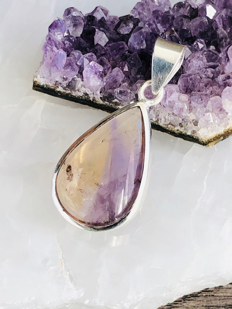 Puede incluir: Un colgante en forma de l&aacute;grima con una piedra de amatista de color amarillo y morado claro engastada en un bisel de plata. El colgante est&aacute; sobre una superficie blanca con un grupo de cristales de amatista en el fondo.