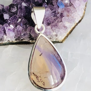 Puede incluir: Un colgante en forma de l&aacute;grima con una piedra clara e iridiscente engastada en un bisel de plata. La piedra tiene un tono p&uacute;rpura claro con vetas m&aacute;s oscuras. El colgante cuelga de una argolla de plata.