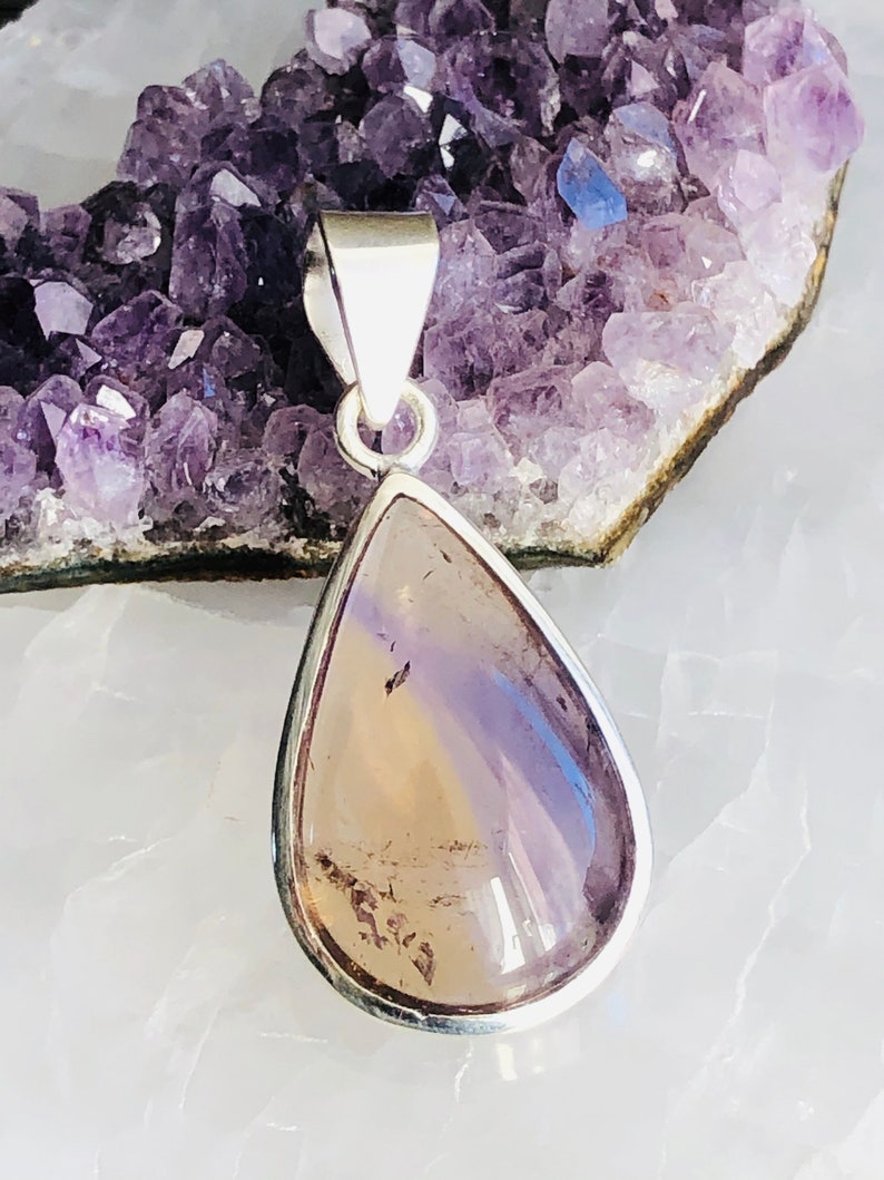 Puede incluir: Un colgante en forma de l&aacute;grima con una piedra clara e iridiscente engastada en un bisel de plata. La piedra tiene un tono p&uacute;rpura claro con vetas m&aacute;s oscuras. El colgante cuelga de una argolla de plata.