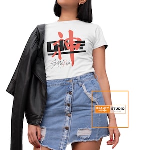 Puede incluir: Camiseta blanca con un gráfico rojo y negro que dice "SKZ" y "Stray Kids" en una fuente estilizada. La persona que lleva la camiseta también lleva una chaqueta de cuero negra y una falda vaquera azul claro.