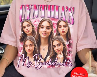 May include: A light pink t-shirt featuring a graphic print with the text "CYNTHIA'S" at the top and "My Bachelorette" at the bottom. The design includes five photographic portraits of the same person arranged over a pink, cloudy background.