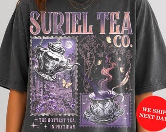May include: A charcoal gray short-sleeve t-shirt featuring a graphic print with the text SURIEL TEA CO. THE HOTTEST TEA IN PRYTHIAN. The design includes a teapot, a teacup with swirling steam, butterflies, and floral elements in purple tones.