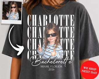 May include: A charcoal gray oversized t-shirt featuring a repeating white text graphic of the name CHARLOTTE. Below the text is a photo of a child wearing sunglasses and a striped shirt, with the words Bachelorette, MIAMI, FLORIDA, 2026 printed below.