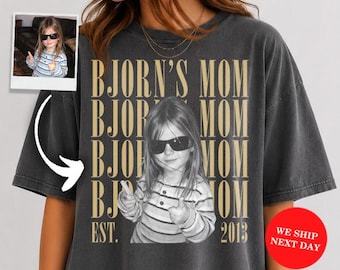 May include: A charcoal gray short-sleeve t-shirt featuring a large graphic print of a child wearing sunglasses and giving a thumbs up. The text "BJORN'S MOM" is repeated in gold lettering behind the child, with "EST. 2013" printed at the bottom.