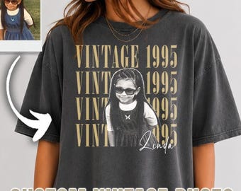 May include: A charcoal gray oversized t-shirt featuring a graphic print of a child wearing sunglasses. The text VINTAGE 1995 is repeated four times in a tan font, with the name Linda written in script below the image. A small inset photo shows the child.