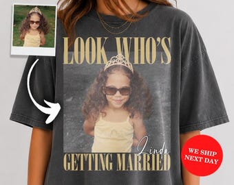 May include: A charcoal gray oversized t-shirt featuring a large graphic print of a child wearing a tiara and sunglasses. The text "LOOK WHO'S GETTING MARRIED" is printed in a beige serif font above and below the central photographic image.