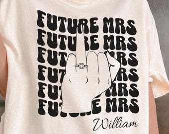 May include: A cream-colored t-shirt featuring a black graphic print of the text "FUTURE MRS" repeated in a wavy font. In the center, a hand illustration shows a middle finger wearing a ring, with the name "William" printed below in script.