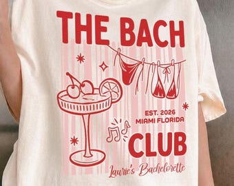 May include: A cream-colored t-shirt featuring red graphic text that reads THE BACH CLUB, EST. 2026 MIAMI FLORIDA, and Laurie's Bachelorette. The design includes a cocktail glass with cherries, a clothesline with swimwear, and musical notes.