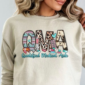 May include: Beige sweatshirt with the letters "OMA" in a colorful design, with the words "Qualified Medical Aide" below. The letters have patterns including a tribal design, leopard print, and cowhide. Medical-themed graphics are included.