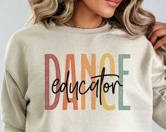 Op de afbeelding: Een crèmekleurige sweatshirt met de tekst "DANCE educator" op de voorkant in een kleurrijk, getextureerd lettertype. Het woord "DANCE" is gestapeld, waarbij elke letter een andere kleur heeft. Het woord "educator" is in een zwart scriptlettertype.