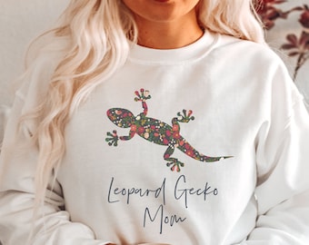 Puede incluir: Sudadera blanca con un diseño de gecko con estampado floral. El texto "Leopard Gecko Mom" está impreso debajo del gecko. La sudadera es de estilo cómodo e informal.
