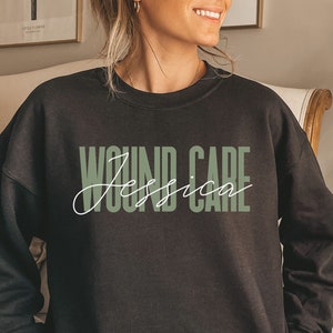 May include: A dark gray sweatshirt with the words "WOUND CARE" in a green block font and the name "Jessica" in a white cursive font. The sweatshirt is a crew neck style.