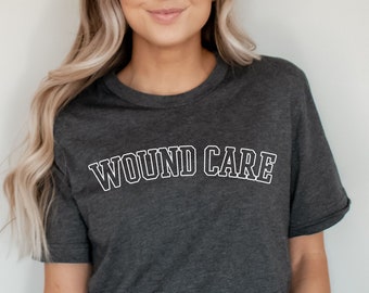 Puede incluir: Una camiseta gris oscuro con el texto "WOUND CARE" impreso en blanco en la parte delantera. El texto está en un estilo de fuente universitario.
