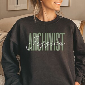 May include: Black sweatshirt with the text "ARCHIVIST Jessica" in green and white lettering.