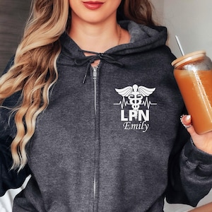 Puede incluir: Una sudadera con capucha gris oscuro con un gráfico blanco de un símbolo de caduceo médico, una línea de latido del corazón y el texto "LPN Emily".