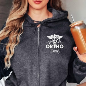 以下が含まれることがあります： 白い医療記号、心拍、そして「ORTHO Emily」というテキストが書かれた白いグラフィックが施された、ダークグレーのジッパー付きフーディーです。