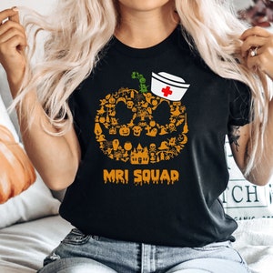 May include: Black t-shirt with a Halloween-themed pumpkin design. The pumpkin is composed of various spooky icons and a nurse's hat. The text "MRI SQUAD" is printed below the pumpkin. A decorative pumpkin pillow is in the background.